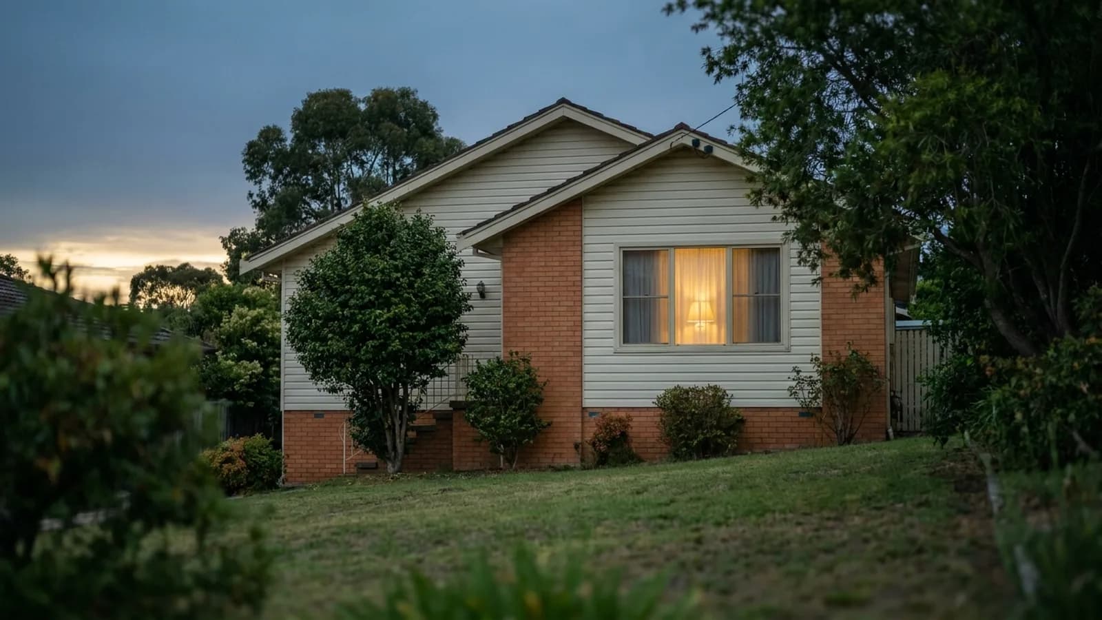 A modest family home viewed across the front lawn at dusk, one interior light on